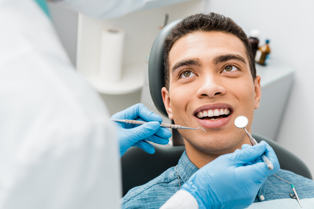 Patient smiling during dental checkup – Ideal Dental Houston TX A young man sits in a dental chair smiling as a dentist wearing gloves uses dental instruments for an oral examination, reflecting comfort and attentive care – Ideal Dental Houston TX