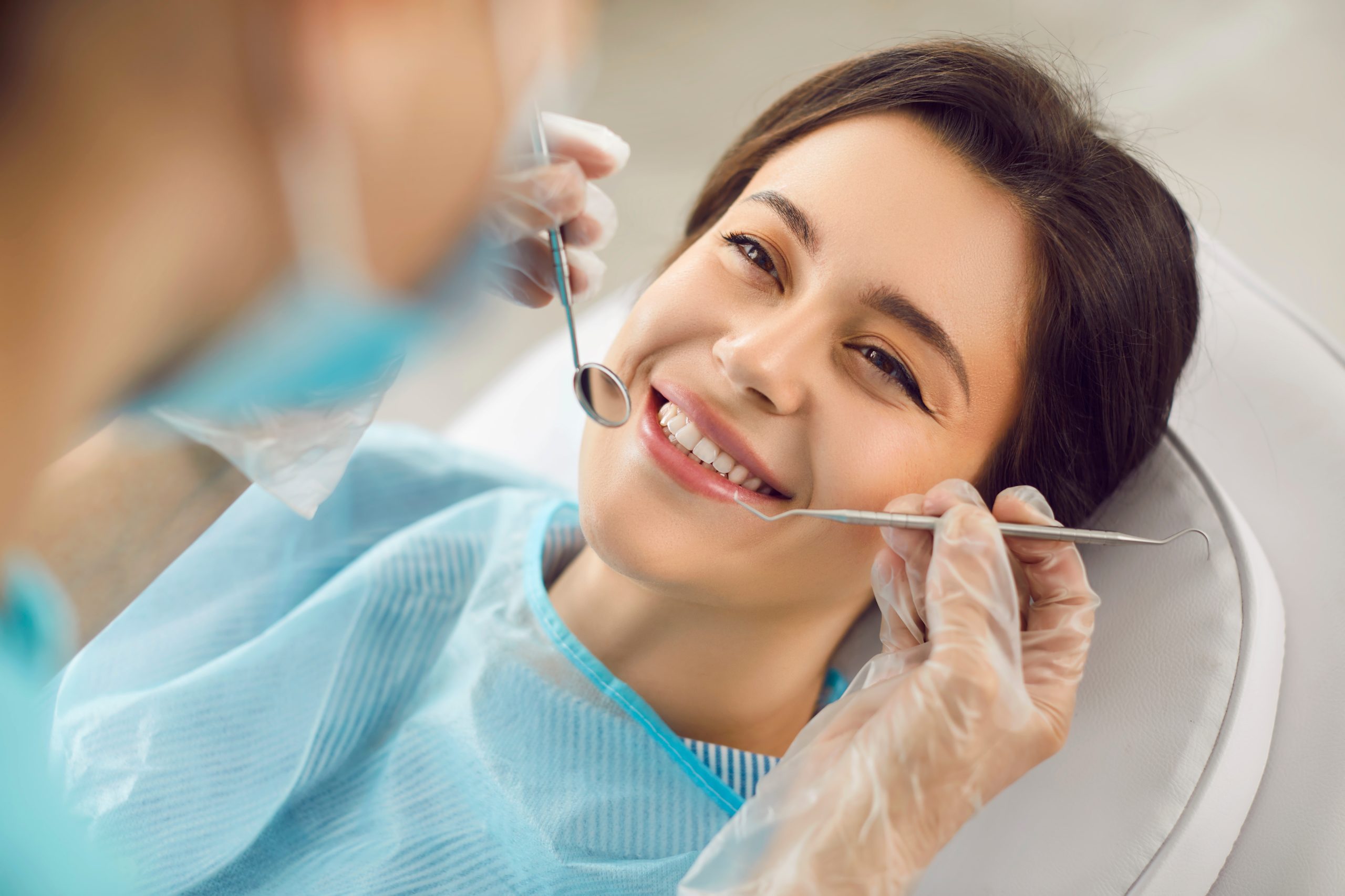 A young woman sits in a dental chair smiling as a dentist wearing gloves uses dental instruments for an oral examination, reflecting comfort and attentive care – Ideal Dental Houston TX